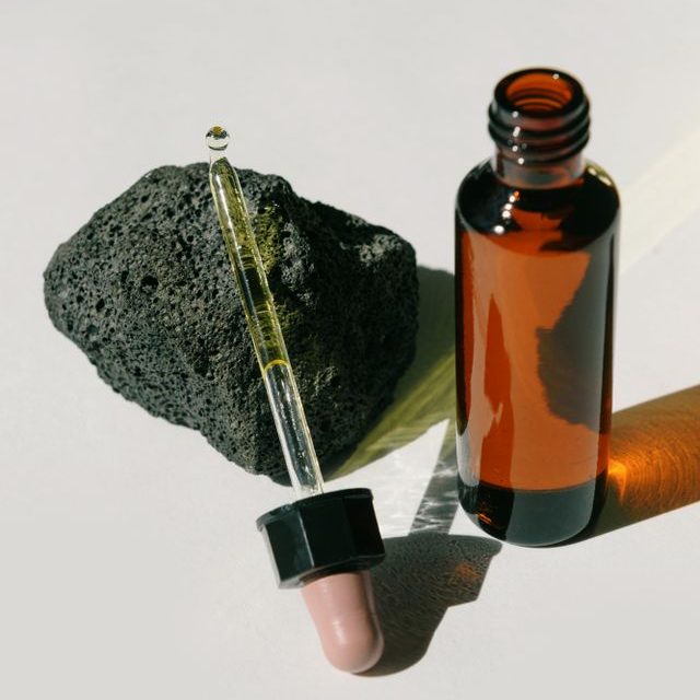 Amber glass dropper bottle and textured lava rock on white background under sunlight.